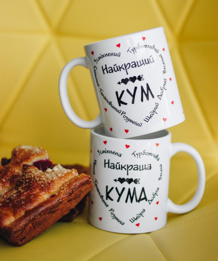 Парні білі чашки з принтами Найкращий кум та Найкраща кума на фоні жовтої стіни.